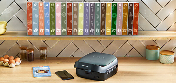 Opiekacz SW862B na blacie obok telefonu i książki. Nad nim półka z kolorowymi książkami.