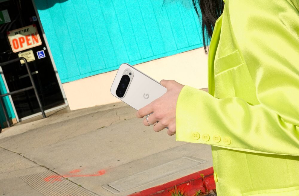 Osoba w żółtej marynarce trzyma biały smartfon z logo \'G\'. W tle widoczny jest budynek z niebieską ścianą i znak \'WE\'RE OPEN\'.