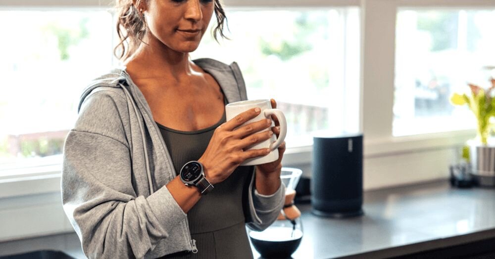 Osoba w sportowym stroju trzyma kubek, na nadgarstku ma szary smartwatch. W tle widać kuchnię z oknem i sprzętem AGD.