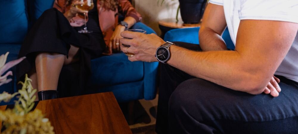 Osoba siedząca na kanapie trzyma w ręku szklankę, na nadgarstku ma szary smartwatch. W tle widać fragment innej osoby oraz niebieską kanapę.