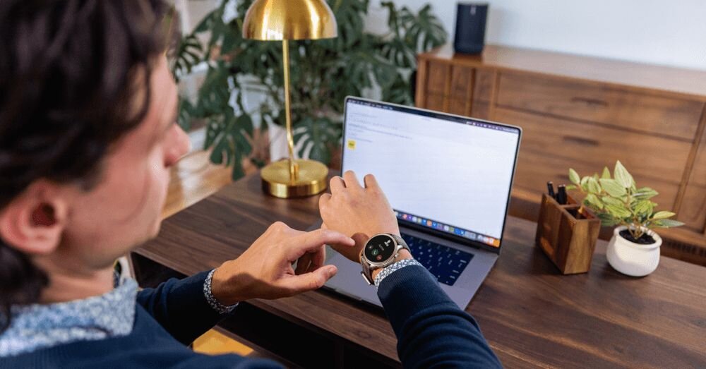 Osoba siedzi przy biurku i patrzy na smartwatch na nadgarstku. Na biurku znajduje się otwarty laptop, lampa i roślina w doniczce.