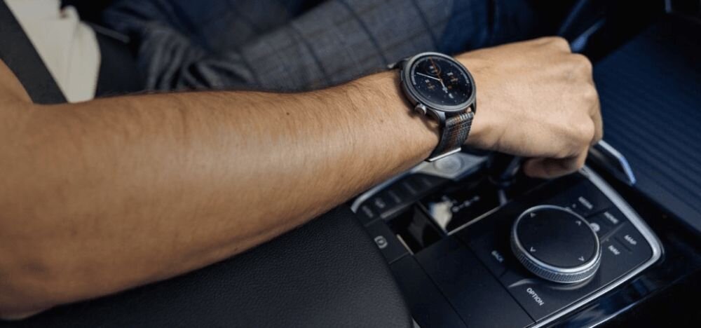 Osoba siedząca w samochodzie ma na nadgarstku szary smartwatch z okrągłą tarczą. W tle widoczna jest konsola samochodowa z pokrętłem i przyciskami.
