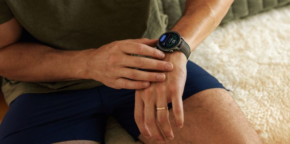 Osoba siedzi na łóżku i nosi szary smartwatch na nadgarstku. Smartwatch wyświetla kolorowy ekran z informacjami.