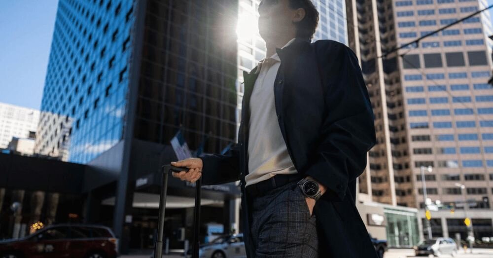 Osoba w ciemnym płaszczu stoi na tle wysokich budynków w mieście, nosząc na nadgarstku smartwatch. W tle widać szklane fasady wieżowców i przejeżdżające samochody.