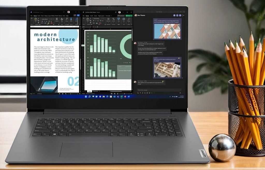 Na biurku stoi otwarty laptop z widocznym ekranem, na którym wyświetlane są różne aplikacje, w tym dokument tekstowy i wykresy. Obok laptopa znajduje się pojemnik z ołówkami i metalowa kula.