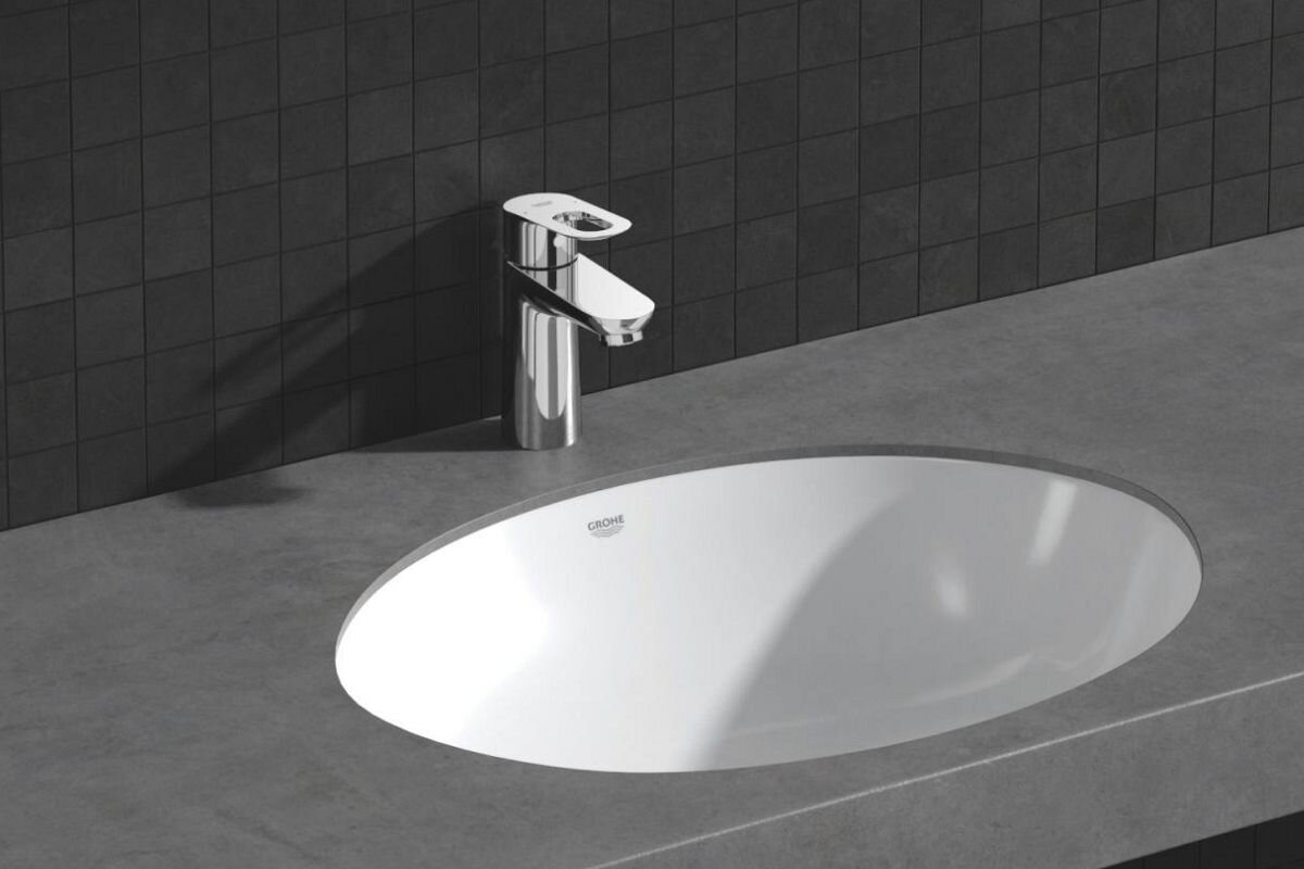 BATERIA UMYWALKOWA GROHE BAULOOP 23337000 CHROM Zdjęcie przedstawia umywalkę nablatową o owalnym kształcie osadzoną w betonowym blacie, z zamontowaną nad nią chromowaną baterią stojącą na tle ciemnej, kwadratowej glazury