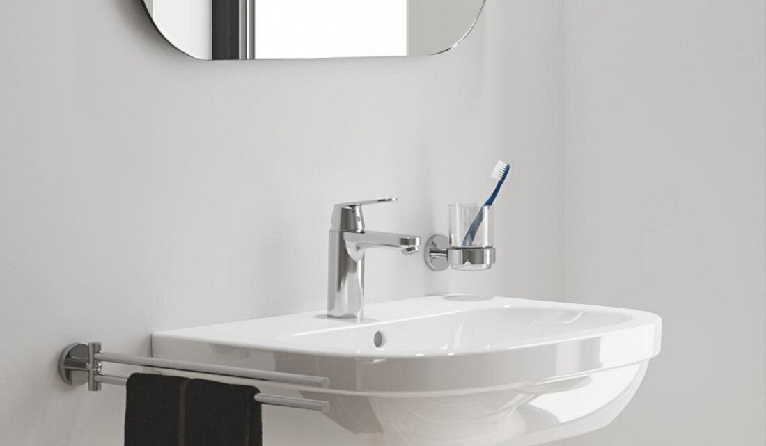 BATERIA UMYWALKOWA GROHE EUROSMART COSMOPOLITAN 23327000 CHROM Zdjęcie przedstawia minimalistyczną umywalkę z prostą, chromowaną baterią oraz szklanym kubkiem na szczoteczkę, zamocowanym na ścianie obok. Całość utrzymana jest w jasnej kolorystyce, podkreślającej czystość i funkcjonalność łazienki
