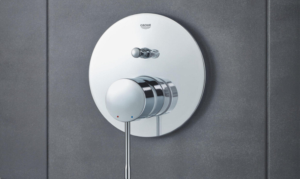 BATERIA WANNOWA PODTYNKOWA GROHE ESSENCE Zdjęcie przedstawia chromowaną baterię prysznicową zamontowaną na ścianie z ciemnych, matowych płytek. Światło podkreśla połysk metalu, tworząc elegancki efekt