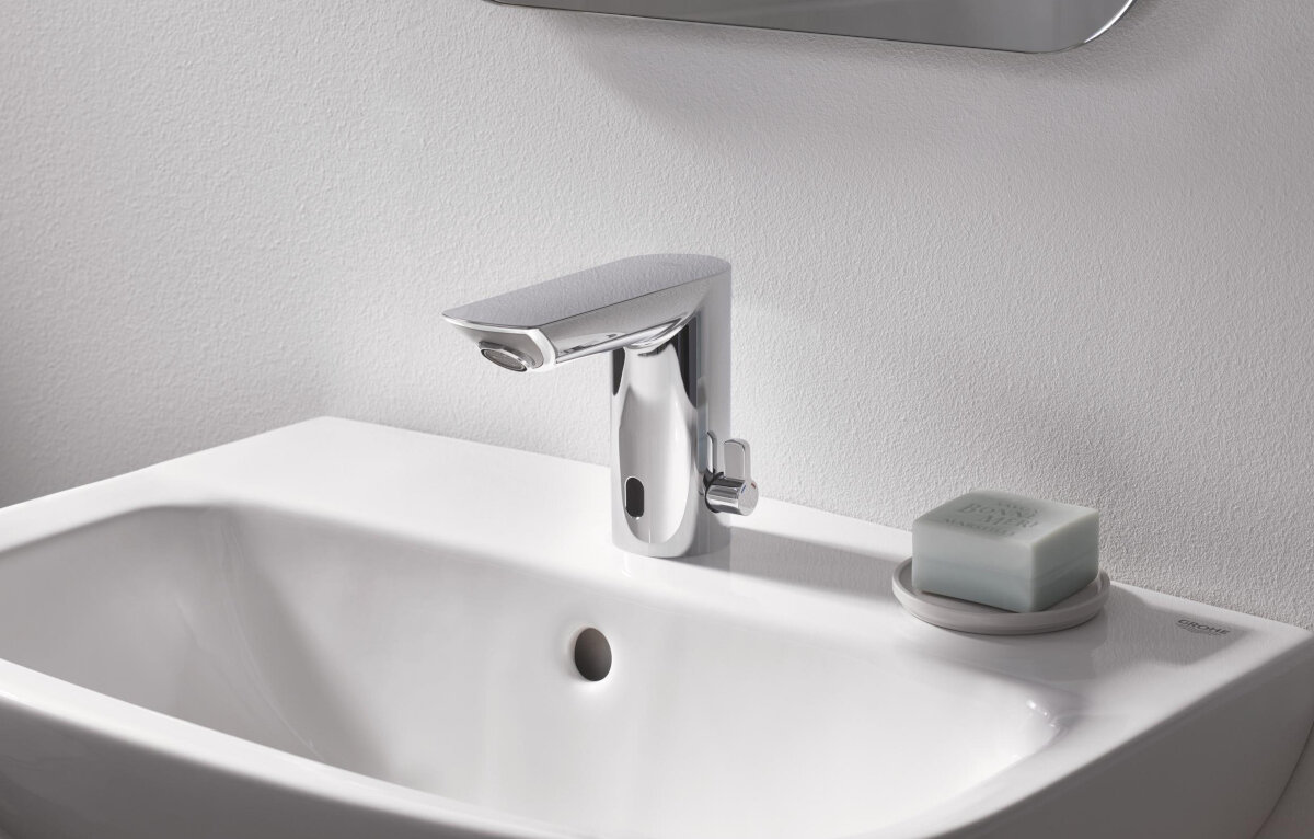 BATERIA UMYWALKOWA GROHE BAU COSMOPOLITAN E ELEKTRONICZNA 36451000 CHROM Na zdjęciu widać chromowaną armaturę bezdotykową zamontowaną na białej umywalce, obok której stoi mydło w kostce na spodku. Ujęcie prezentuje baterię w realistycznym kontekście łazienkowym, podkreślając jej wygląd