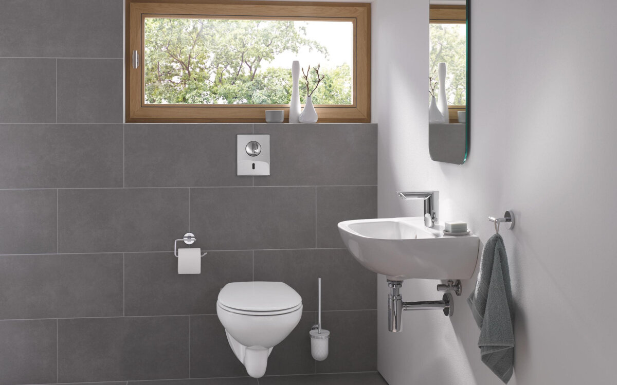 BATERIA UMYWALKOWA GROHE BAU COSMOPOLITAN E ELEKTRONICZNA 36451000 CHROM Zdjęcie ukazuje minimalistyczną łazienkę z zawieszoną toaletą, umywalką i chromowaną armaturą oraz oknem wpuszczającym naturalne światło. Na ścianie znajdują się proste akcesoria, dzięki czemu wnętrze wygląda czysto i elegancko regulacja temperatury wody uchwyt boczny armatury chromowana powierzchnia armatury higiena w łazience mniej bakterii armatura publiczna