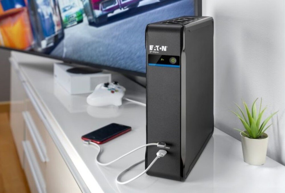 Na białej szafce stoi czarny zasilacz UPS z logo \'EATON\' i podłączonymi kablami USB. Obok znajduje się smartfon, kontroler do konsoli i mała roślina w doniczce, a w tle widać ekran telewizora.