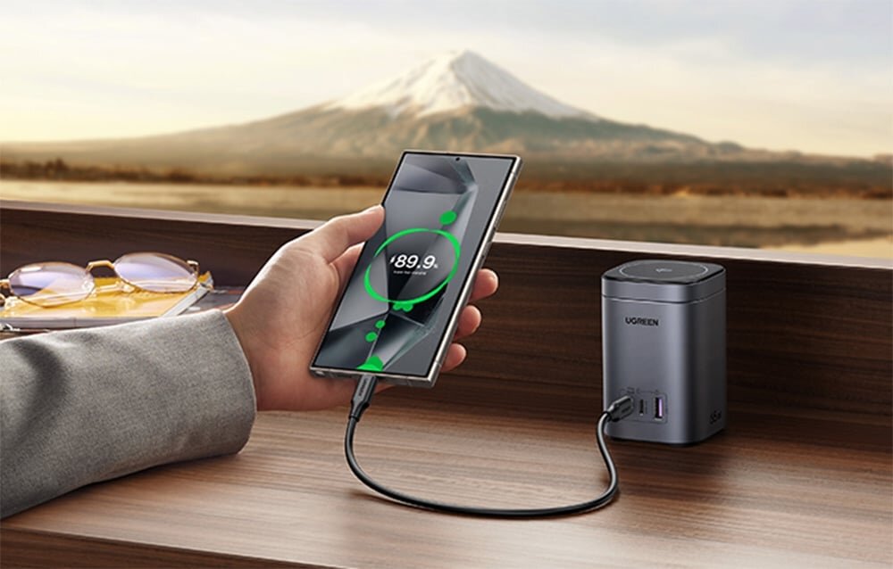 Osoba trzyma smartfon z widocznym wskaźnikiem ładowania na ekranie, podłączony do ładowarki Ugreen z dwoma portami USB-C i jednym USB. W tle widoczny jest krajobraz z górą oraz okulary przeciwsłoneczne na drewnianej powierzchni.