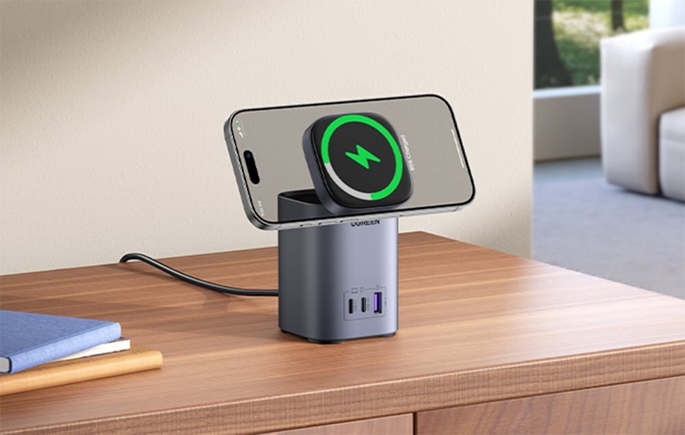 Na drewnianym blacie stoi ładowarka z telefonem umieszczonym na górze, z widocznym symbolem ładowania. Ładowarka ma dwa porty USB-C i jeden USB, a na obudowie widnieje napis \'UGREEN\'.