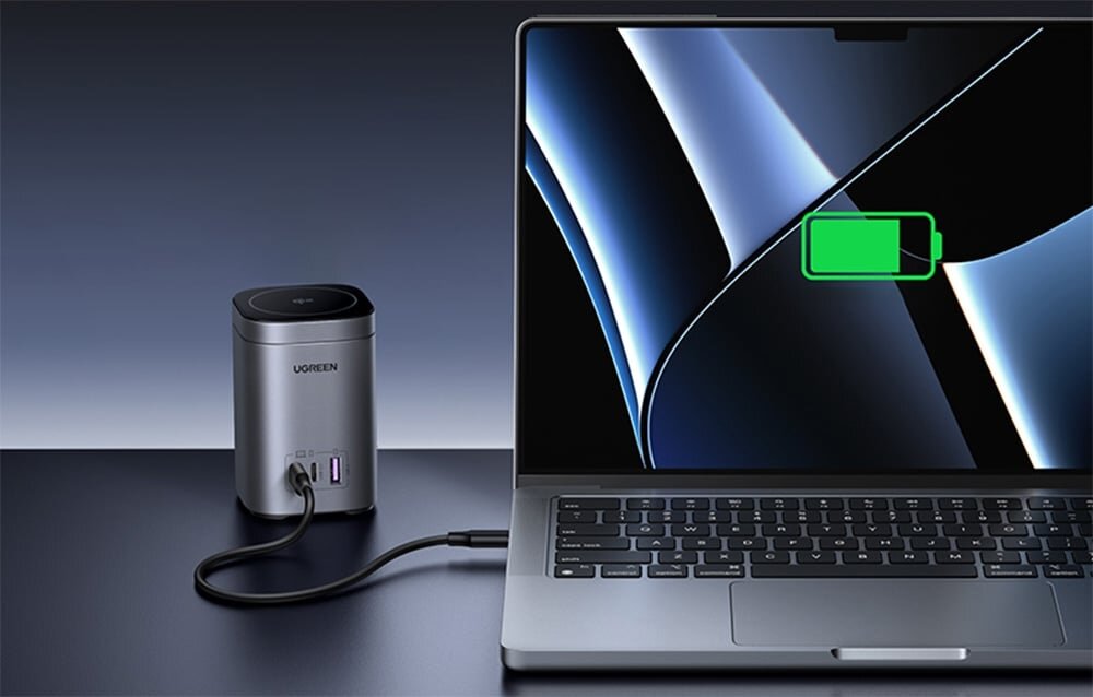 Na obrazie widać laptopa z zieloną ikoną baterii na ekranie, podłączonego do ładowarki z logo UGREEN. Ładowarka ma dwa porty USB-C i jeden port USB, a całość znajduje się na ciemnym tle.