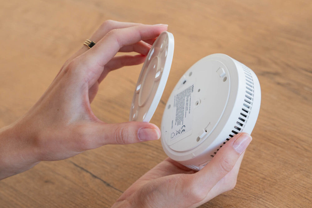 CZUJNIK DYMU I TLENKU WĘGLA VÖGEL VCA200 Dwie dłonie trzymają rozdzielone elementy czujnika dymu i tlenku węgla (czadu) VÖGEL nad drewnianym blatem. Jedna dłoń trzyma okrągłą płytę montażową, a druga główną część czujnika, na której widoczne są kratki wentylacyjne i naklejka informacyjna.