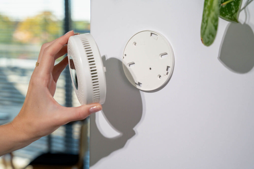 CZUJNIK DYMU I TLENKU WĘGLA VÖGEL VCA200 Detal dłoni z pierścionkiem trzymającej okrągły czujnik dymu i tlenku węgla (czadu) VÖGEL. Czujnik jest odłączany od okrągłej płyty montażowej przymocowanej do białej ściany. W prawym górnym rogu fragment zielonej rośliny, w tle okno.