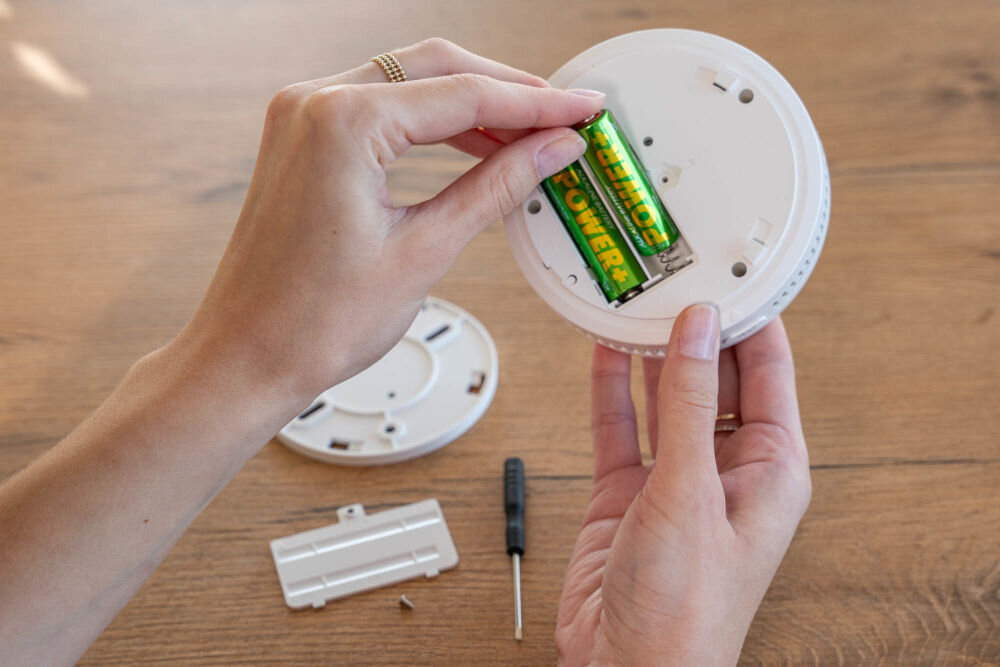 CZUJNIK DYMU I TLENKU WĘGLA VÖGEL VCA200 Dwie dłonie instalują lub wymieniają dwie baterie (zielone z napisem POWER) w komorze baterii znajdującej się w tylnej części czujnika dymu i tlenku węgla (czadu) VÖGEL. Obok na drewnianym blacie leży mały śrubokręt, pokrywa komory baterii i płyta montażowa.