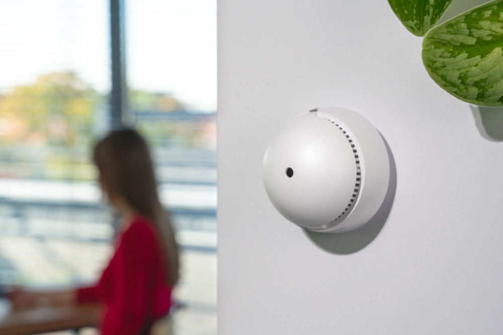 Czujnik dymu VÖGEL VSA200 Miniaturowy, biały czujnik dymu VÖGEL VSA200 zamontowany na białej ścianie w pomieszczeniu biurowym. W tle, rozmyta postać kobiety siedzącej przy biurku i zielona roślina.