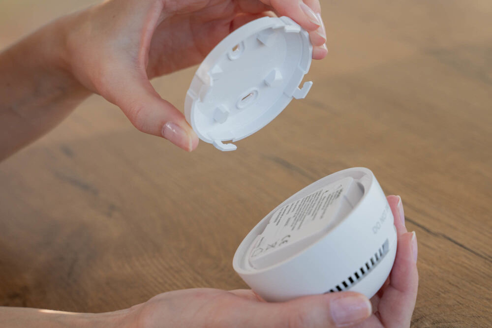 Czujnik dymu VÖGEL VSA200 Ujęcie przedstawiające dwie dłonie. Jedna trzyma główną część czujnika dymu VÖGEL VSA200 (część z elektroniką i baterią), a druga trzyma białą płytę montażową. Elementy są rozdzielone, ukazując sposób montażu.