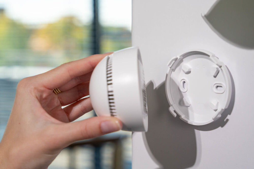 Czujnik dymu VÖGEL VSA200 Detal dłoni trzymającej główne urządzenie czujnika dymu VÖGEL VSA200. Obok widoczna jest biała płyta montażowa, która jest przytwierdzona do ściany.