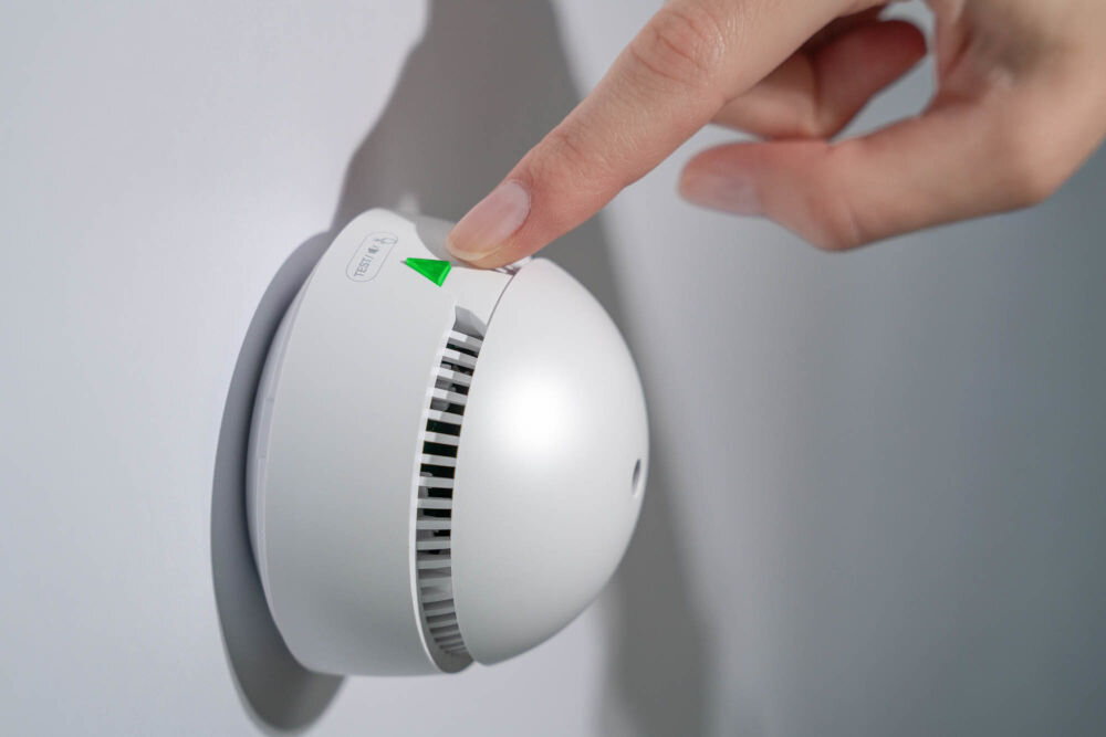 Czujnik dymu VÖGEL VSA200 Ręka naciskająca przycisk testu/wyciszenia (zielony trójkąt ze znakiem dłoni) na zamontowanym na ścianie białym, miniaturowym czujniku dymu VÖGEL VSA200