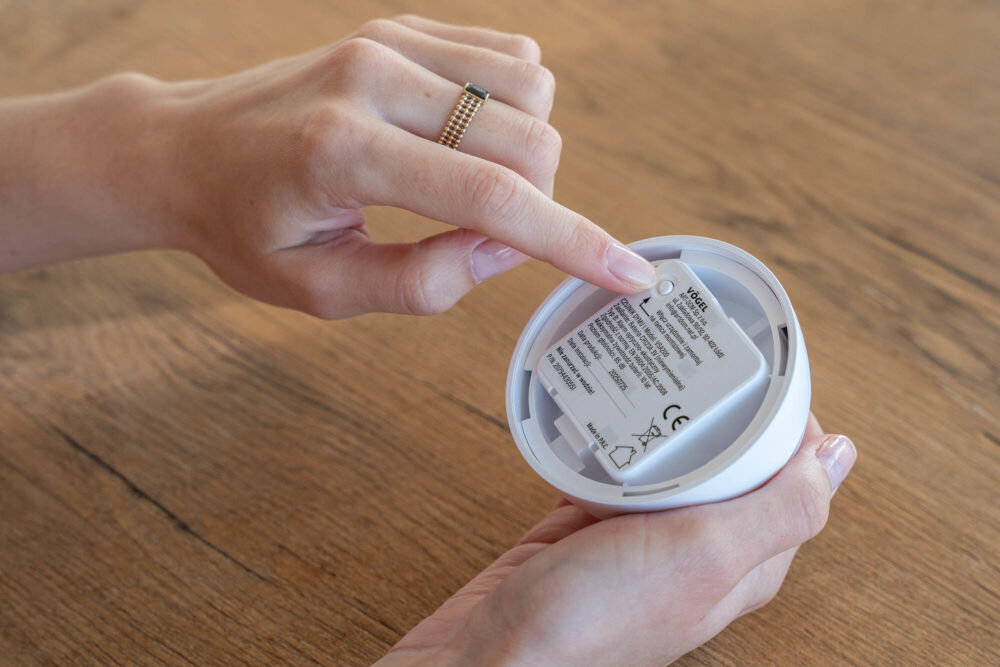 Czujnik dymu VÖGEL VSA200 Dłoń wskazująca na naklejkę z informacjami technicznymi i certyfikatami (CE) umieszczoną na spodzie miniaturowego czujnika dymu VÖGEL VSA200. Czujnik trzymany jest nad drewnianym blatem.