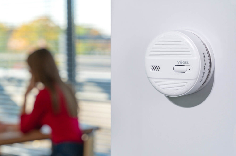 Czujnik dymu VÖGEL VSA100 Okrągły, biały czujnik dymu VÖGEL VSA100 zamontowany na jasnej ścianie. Na jego boku widoczna jest inskrypcja Wymienić 10 lat po instalacji. W tle po lewej stronie, rozmyta postać kobiety w czerwonej koszulce siedzącej przy oknie