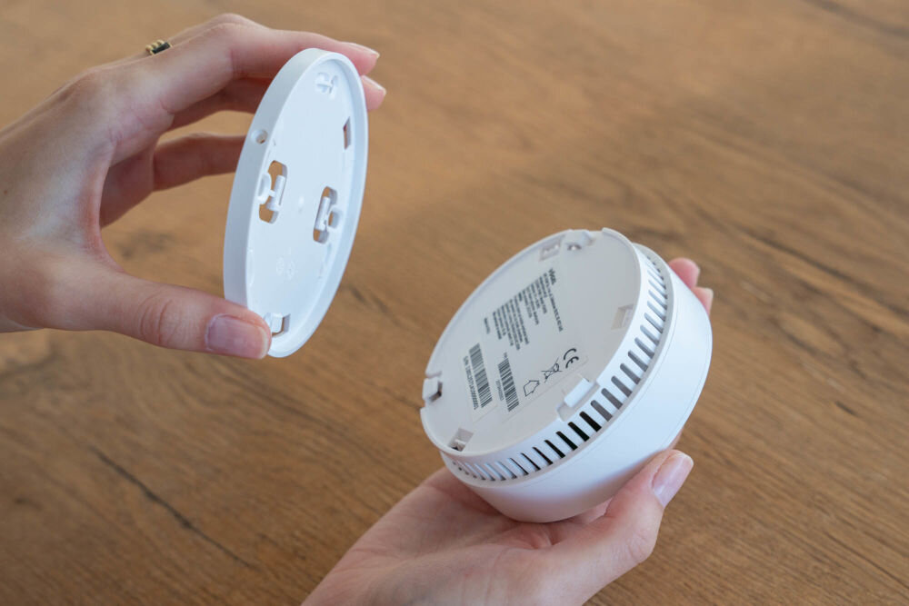 Czujnik dymu VÖGEL VSA100 Dwie dłonie trzymają rozdzielone elementy czujnika dymu VÖGEL VSA100 nad drewnianym blatem. Jedna dłoń trzyma okrągłą płytę montażową, a druga główną część czujnika, na której widoczne są kratki wentylacyjne, kod kreskowy i oznaczenia certyfikacyjne CE.