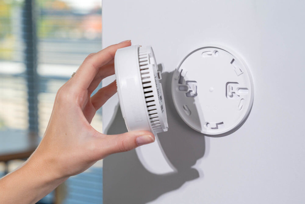 Czujnik dymu VÖGEL VSA100 Detal dłoni trzymającej główną część czujnika dymu VÖGEL VSA100, który jest odłączany od białej płyty montażowej przykręconej do ściany. W tle widoczne okno z widokiem na zewnątrz.