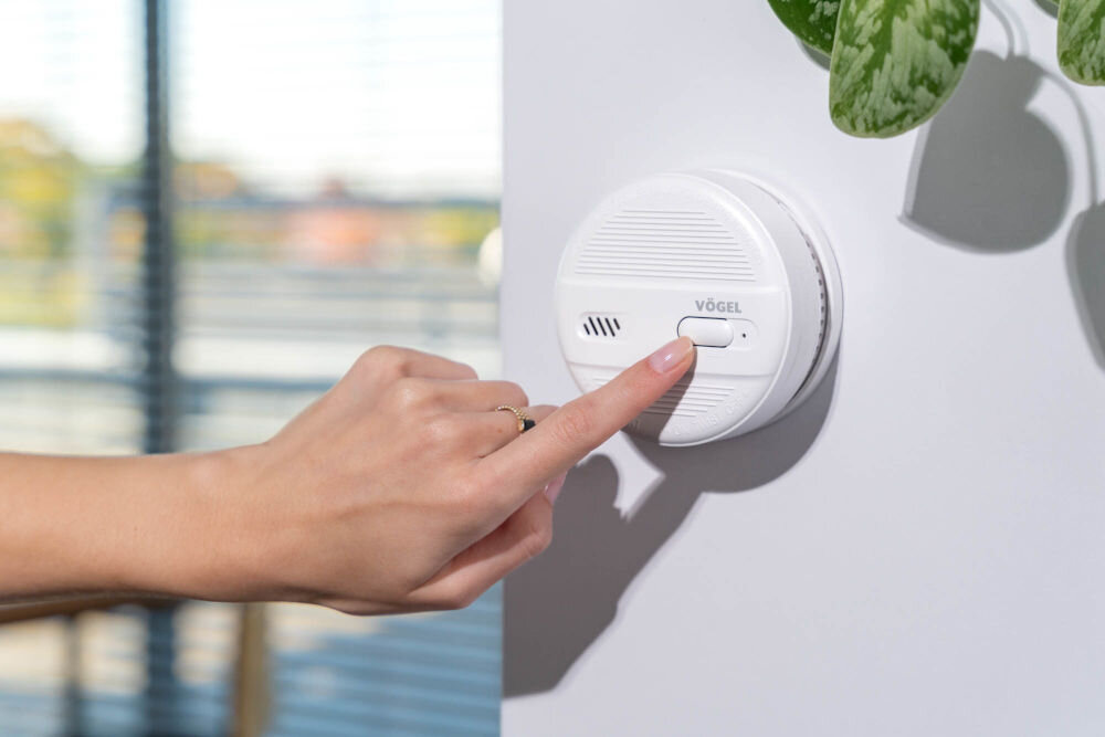 Czujnik dymu VÖGEL VSA100 Ręka osoby z pierścionkiem naciska podłużny przycisk testu na okrągłym, białym czujniku dymu VÖGEL VSA100 zamontowanym na ścianie. W tle, na lewo, rozmyte okno z żaluzjami i element rośliny na górze, po prawej.