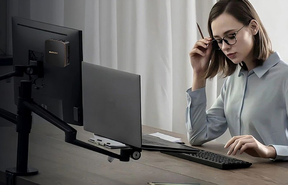 Komputer BLACKVIEW MP20 Kobieta w okularach siedzi przy biurku z klawiaturą i monitorem, do którego z tyłu przymocowany jest mały komputer, mocowanie VESA