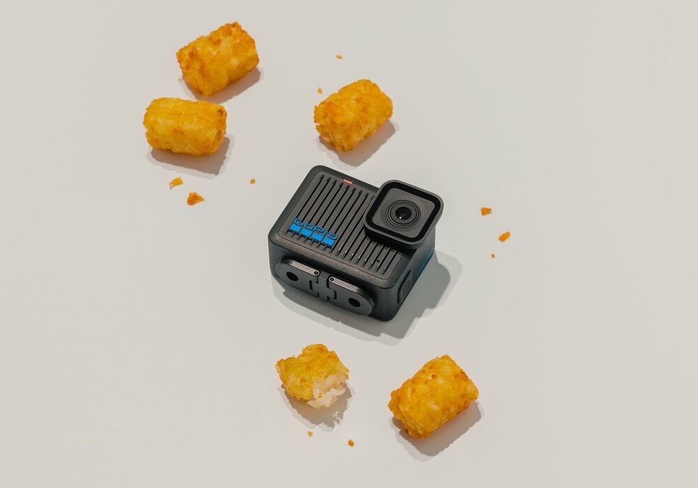 Na białym tle znajduje się czarna kamera sportowa z widocznym obiektywem i niebieskim logo. Wokół kamery leży kilka złocistych krokietów ziemniaczanych.