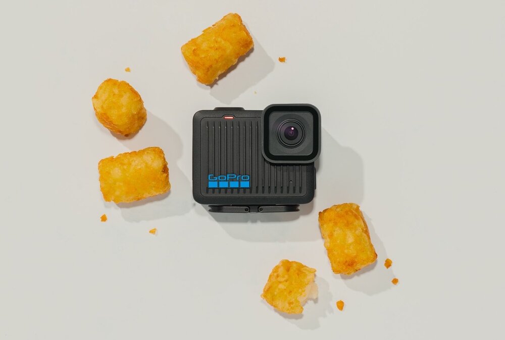 Na białym tle znajduje się czarna kamera sportowa z widocznym obiektywem i logo \'GoPro\' w kolorze niebieskim. Wokół kamery rozmieszczone są kawałki smażonych ziemniaków.