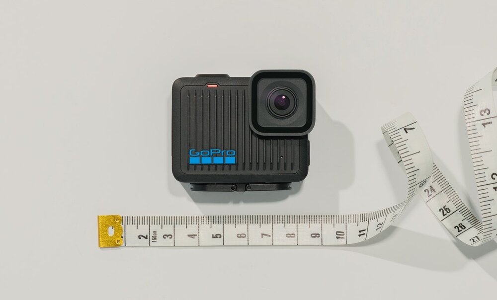 Na obrazie widoczna jest czarna kamera sportowa z logo \'GoPro\' na obudowie. Obok kamery znajduje się rozwinięta miarka z widocznymi centymetrami.