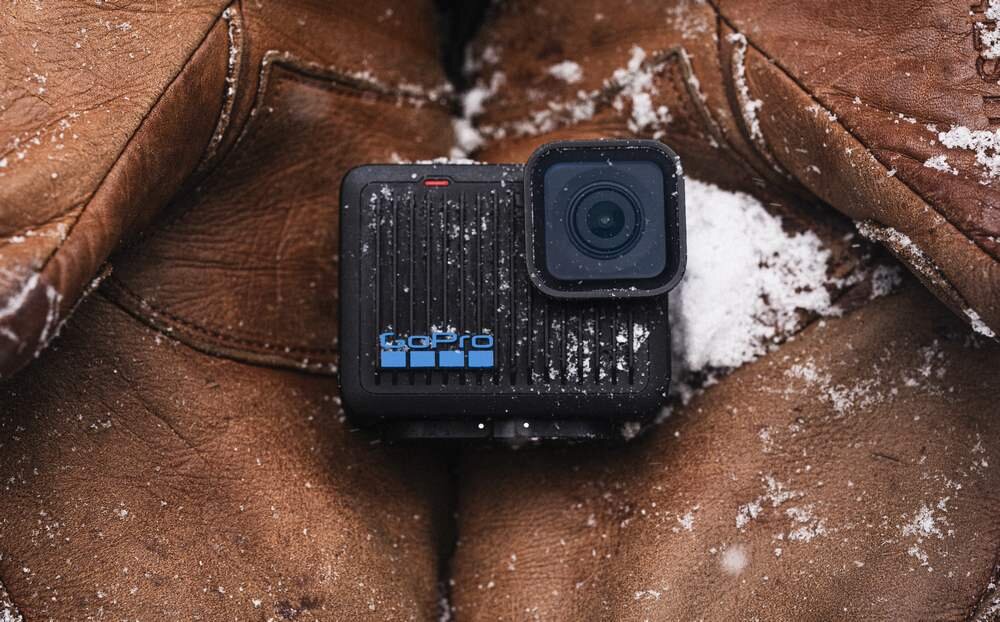 Kamera sportowa z logo \'GoPro\' leży na skórzanych rękawicach pokrytych śniegiem. Kamera ma czarną obudowę i widoczny obiektyw.