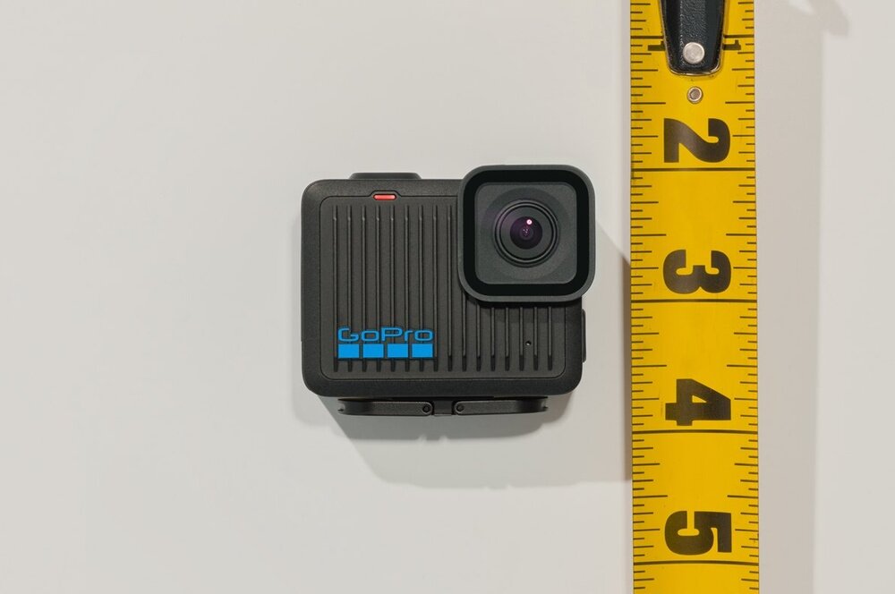 Na białym tle znajduje się czarna kamera sportowa z logo \'GoPro\' w kolorze niebieskim. Obok kamery widoczna jest żółta miarka z oznaczeniami liczbowymi od 1 do 5.