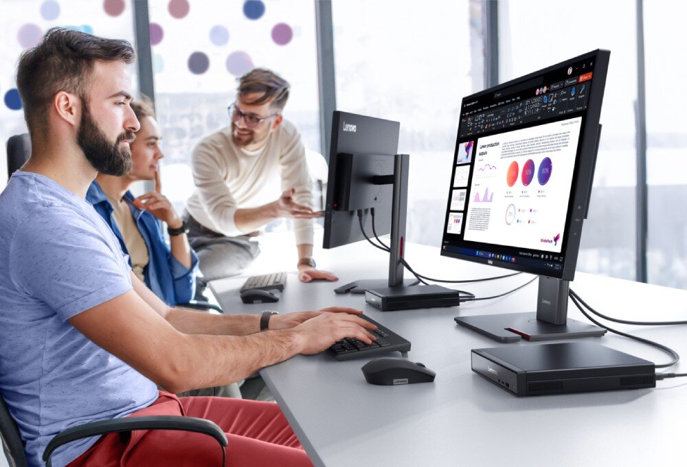 Trzy osoby siedzą przy biurku z komputerami, na których widoczne są wykresy i dokumenty. Na monitorze widnieje logo \'Lenovo\'.