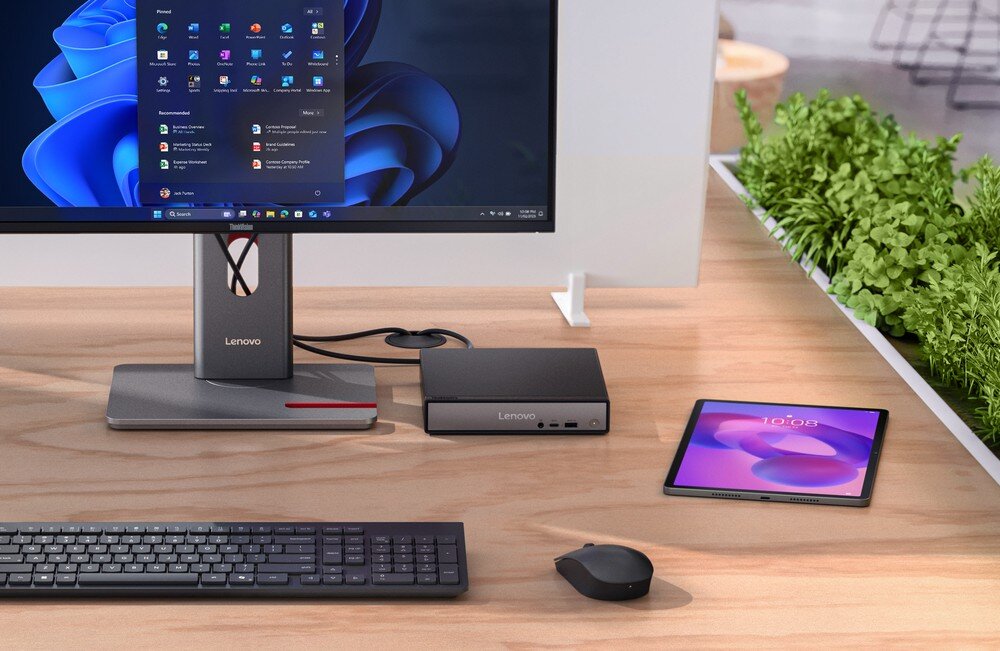 Na biurku znajduje się monitor z logo Lenovo, obok którego stoi kompaktowy komputer Lenovo. Przed monitorem leży klawiatura i myszka, a obok tabletu wyświetla się godzina 10:03.