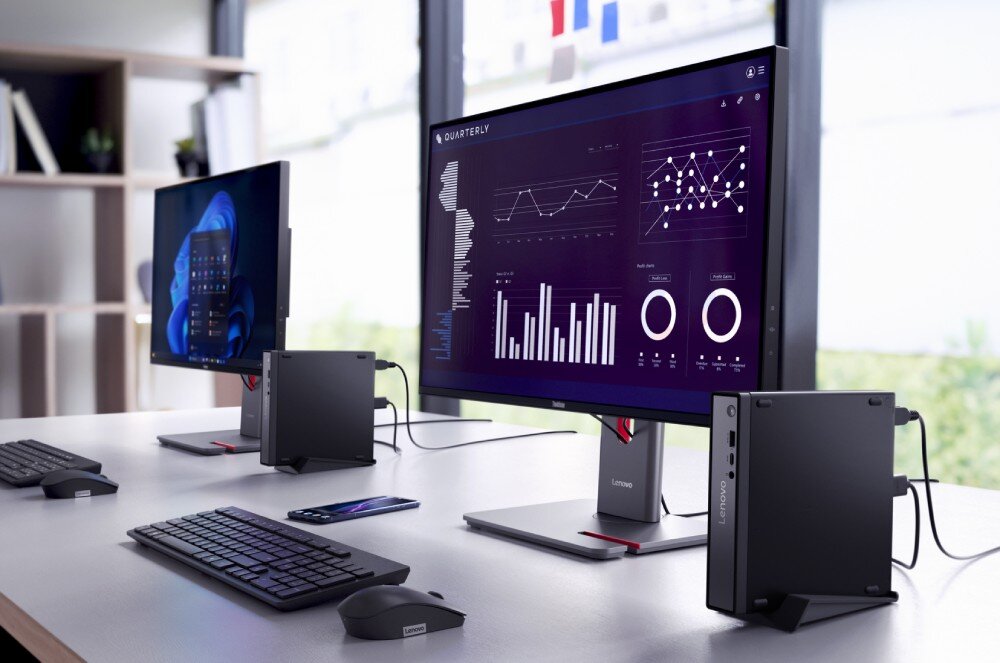 Na biurku znajdują się dwa monitory, klawiatury i myszki, a obok nich stoją komputery Lenovo. Na ekranach widoczne są wykresy i dane, a na jednym z nich napis \'QUARTERLY\'.