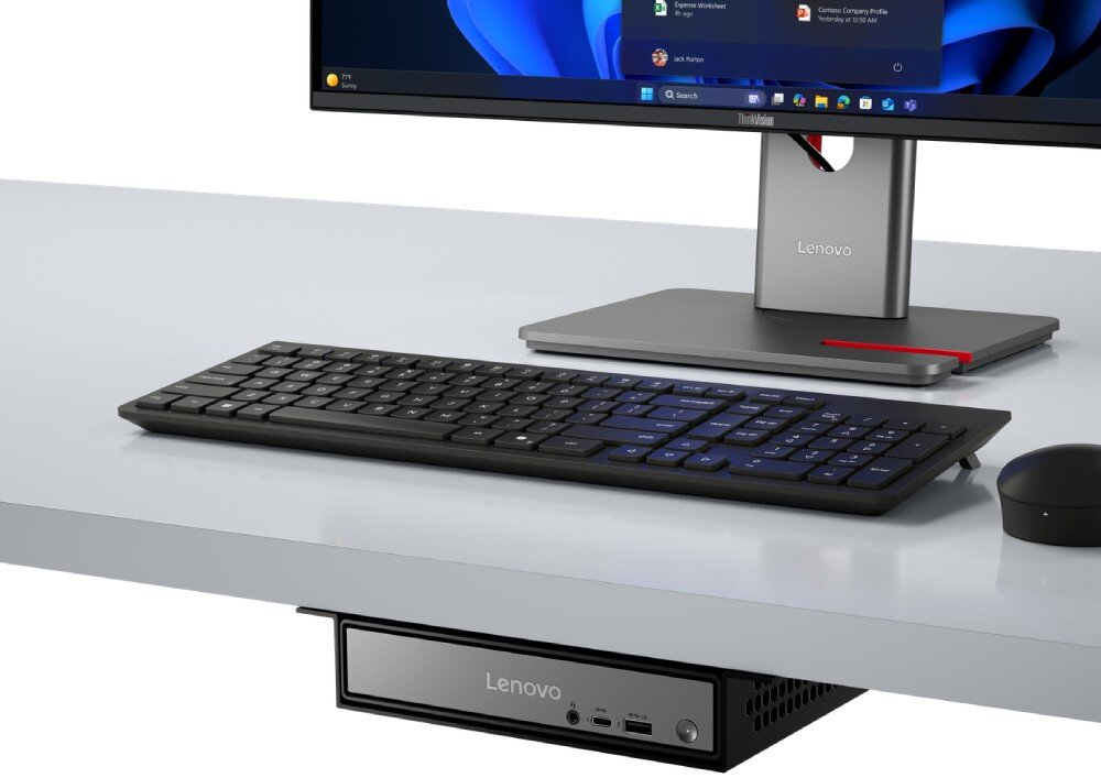 Na biurku znajduje się monitor z logo Lenovo, klawiatura, mysz oraz komputer Lenovo umieszczony pod blatem. Na ekranie widoczny jest interfejs systemu Windows 11.