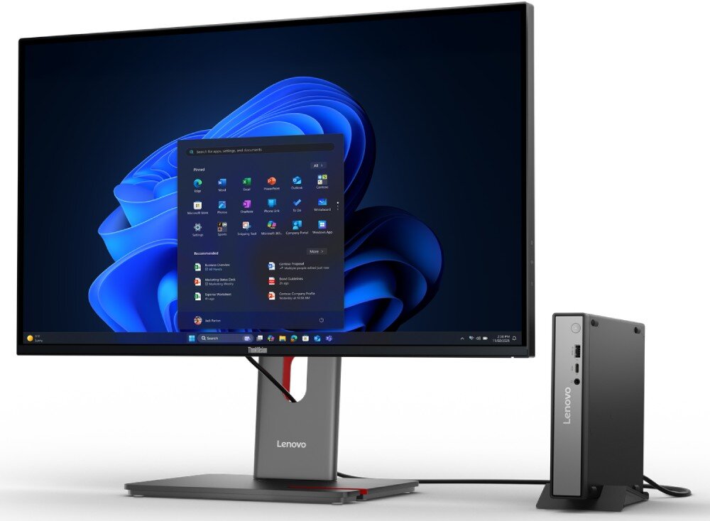 Na obrazie widoczny jest monitor z logo Lenovo, na którym wyświetlany jest ekran startowy systemu Windows 11. Obok monitora znajduje się kompaktowy komputer z napisem Lenovo na obudowie.