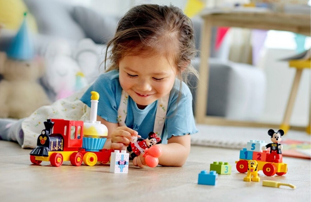 Dziecko bawi się zestawem klocków LEGO z motywem pociągu i postaciami Myszki Miki i Minnie. Na podłodze widoczne są kolorowe klocki i figurki.