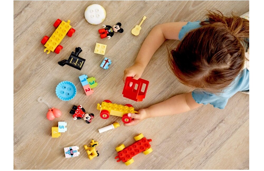 Na podłodze leżą klocki LEGO, w tym figurki Myszki Miki i Minnie oraz elementy pociągu w kolorach czerwonym i żółtym. Dziecko bawi się jednym z elementów pociągu.