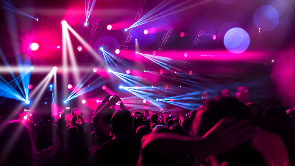 Tłum ludzi na koncercie z kolorowymi światłami w tle, dominują różowe i niebieskie reflektory. Widać unoszące się ręce i telefony wśród publiczności.