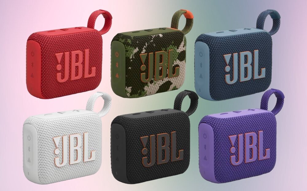 Na obrazie znajduje się sześć głośników mobilnych w różnych kolorach: czerwonym, zielonym z wzorem moro, niebieskim, białym, czarnym i fioletowym. Każdy głośnik ma widoczne logo \'JBL\' oraz pętlę do przenoszenia.