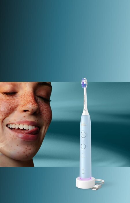 Zbliżenie na twarz uśmiechniętej kobiety, która językiem przesuwa po zębach. Obok szczoteczka soniczna.