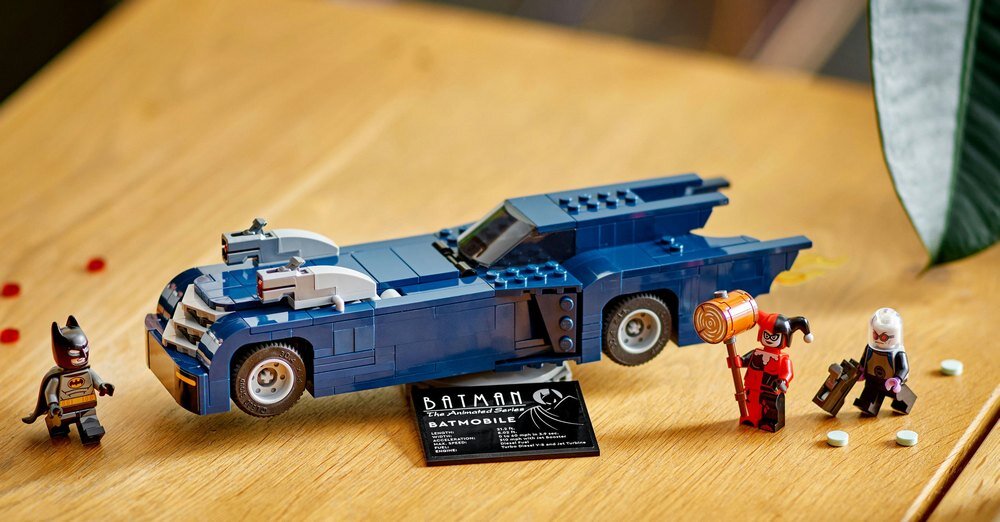 Na drewnianym stole znajduje się model Batmobilu z klocków LEGO, obok którego stoją trzy figurki postaci. Przed modelem umieszczona jest tabliczka z napisem \'BATMAN The Animated Series BATMOBILE\'.