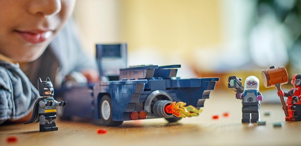 Na drewnianym stole znajduje się zestaw klocków LEGO przedstawiający Batmobil z płomieniem z rury wydechowej oraz trzy figurki: Batman, postać z młotem i postać z niebieską maską. W tle widoczna jest twarz dziecka obserwującego zabawkę.