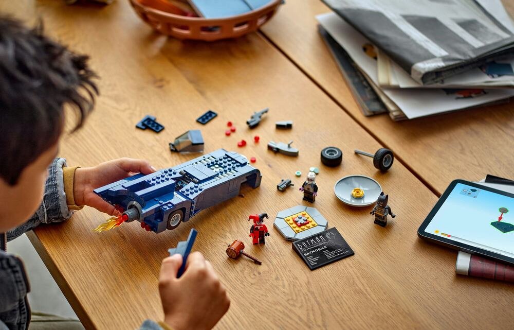Na drewnianym stole znajduje się zestaw klocków LEGO, w tym pojazd i kilka figurek. Obok leży tablet z wyświetlonym obrazem klocków oraz karta z napisem \'BATMAN\'.