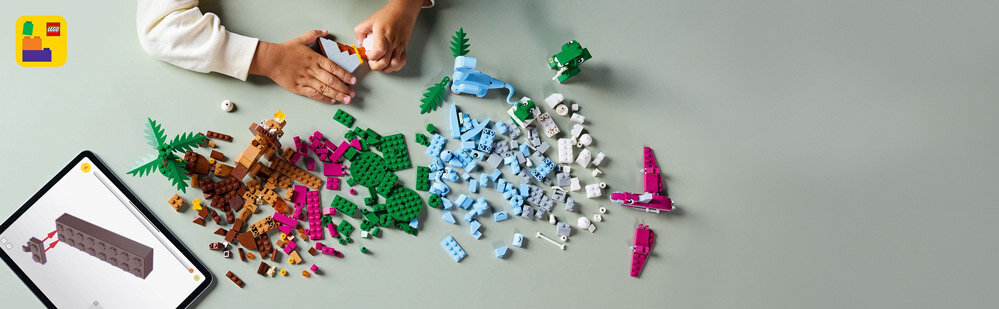 Na stole leżą różnokolorowe klocki LEGO, a obok nich widoczne są ręce osoby składającej model. W lewym górnym rogu znajduje się tablet z wyświetlonym modelem z klocków.